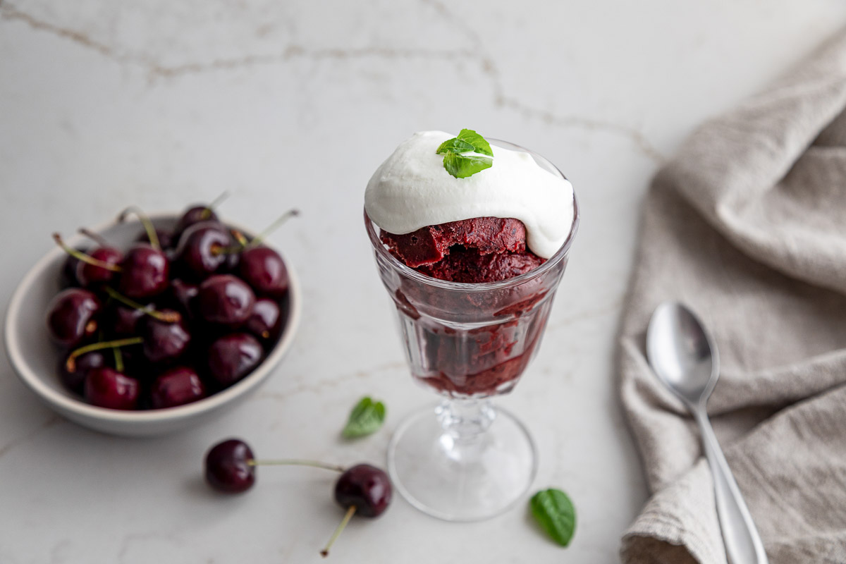 Cherry Sorbet in goblet with basil whipped cream and mint leaf near a bowl of fresh cherries - Dairy Free Sorbet