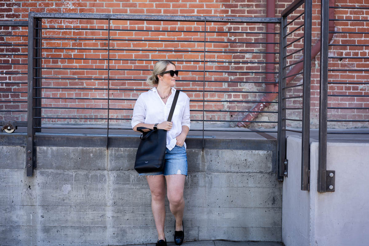 Luci in White Button Down Shirt, Blue Shorts, Black Loafers, & Black crossbody, leaning against wall/fence