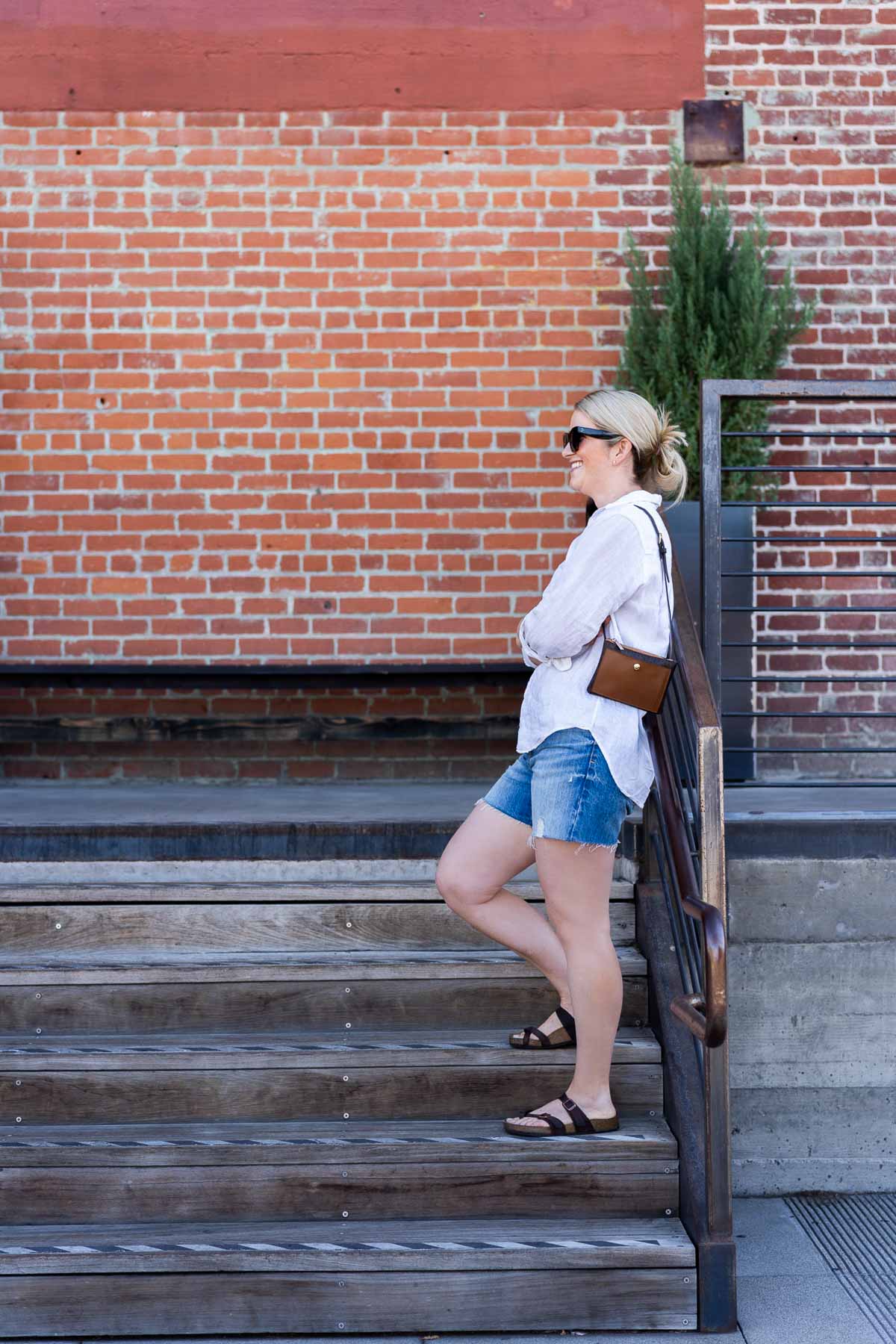Luci in White Button Down Shirt, Blue Shorts, Birkenstock Sandals, & Black crossbody, leaning against Stare Rail